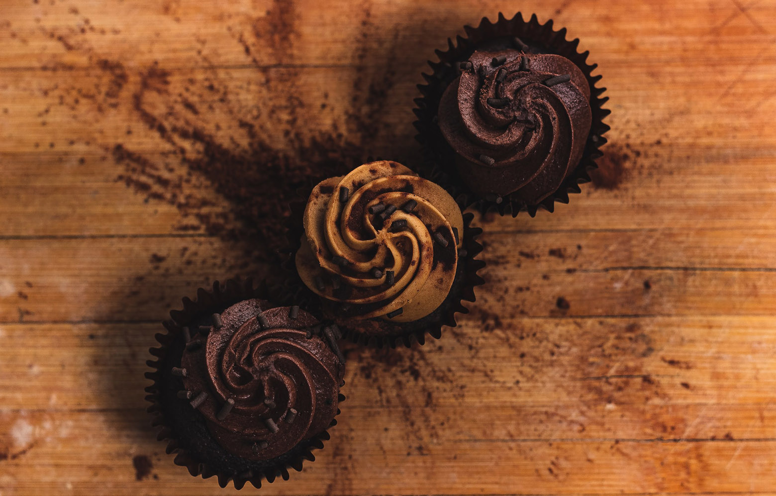Three chocolate cupcakes lined up diagonally on the brown wooden surface. The middle cupcake is light brown, other two are dark brown.