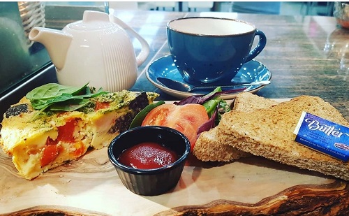 Blu cup, white tea pot, toast with butter packaging, tomato sauce, tomato and quiche on a countertop